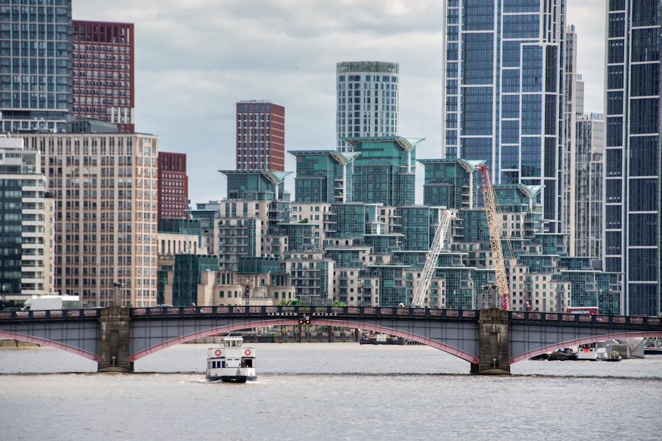 A cityscape featuring modern high-rise buildings with glass facades and varying architectural styles, set against a cloudy sky. In the foreground, a river flows beneath a decorative arched bridge made