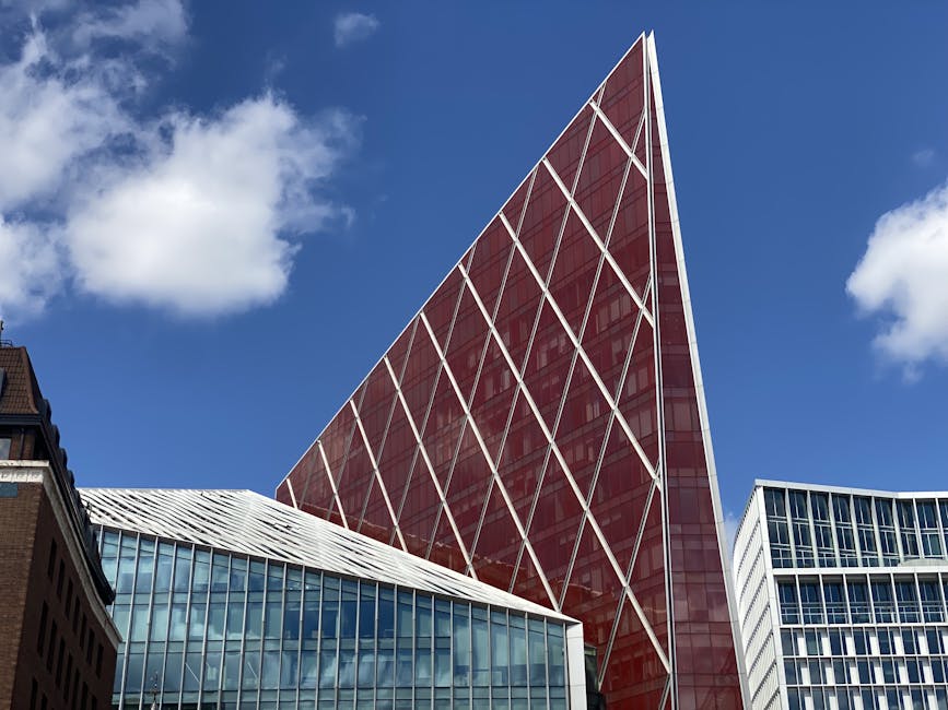 A modern cityscape featuring a prominent red and white glass skyscraper with a crisscross metal framework, contrasted against a bright blue sky with scattered white clouds. In the foreground, parts of